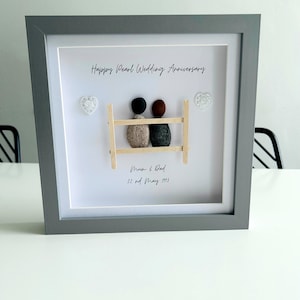 May include: A framed pebble art piece with a gray frame and a white background. The art depicts two figures made of pebbles, sitting on a wooden bench. The text "Happy Pearl Wedding Anniversary" is written at the top of the frame. The text "Mum & Dad 22nd May 1993" is written below the figures.