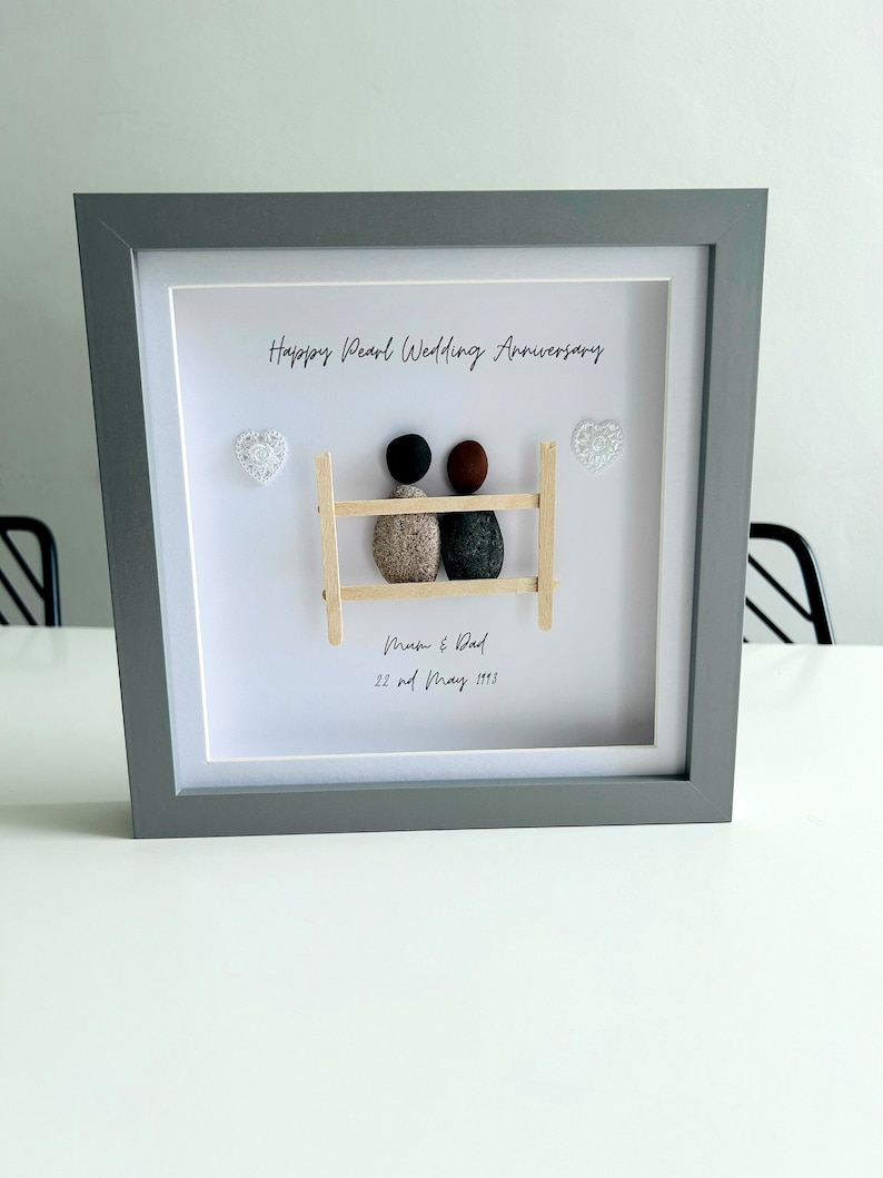 May include: A framed pebble art piece with a gray frame and a white background. The art depicts two figures made of pebbles, sitting on a wooden bench. The text "Happy Pearl Wedding Anniversary" is written at the top of the frame. The text "Mum & Dad 22nd May 1993" is written below the figures.