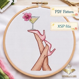 May include: A cross-stitch pattern in a wooden embroidery hoop featuring a martini glass with a pink hibiscus flower, balanced on a pink high-heeled shoe worn on a leg. The design includes text labels reading PDF Pattern and XSP file.