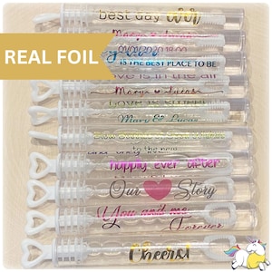May include: A set of ten clear plastic bubble wands with gold and pink foil lettering. The wands are decorated with various wedding-themed messages, including "Best Day Ever", "Love is in the Air", "Our Story", and "Cheers!"