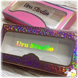 May include: Two eyelash boxes, one pink and one holographic, are displayed. The pink box has a clear window and the text "Uru Studio" in black. The holographic box has a clear window and the text "Uru Studio" in a rainbow gradient.