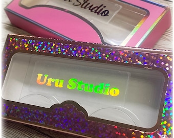 May include: Two eyelash boxes, one pink and one holographic, are displayed. The pink box has a clear window and the text "Uru Studio" in black. The holographic box has a clear window and the text "Uru Studio" in a rainbow gradient.