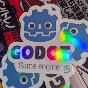 May include: A stack of stickers featuring the "GODOT Game engine" logo in a rainbow holographic design. The stickers have a blue gear-shaped design with the text "GODOT Game engine" in white. The background includes Japanese text and car illustrations.