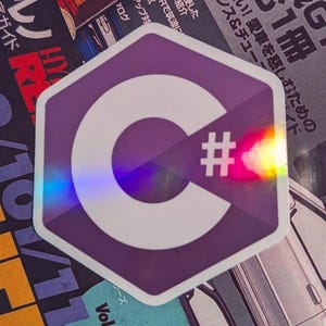 May include: A purple, hexagonal sticker featuring a white "C" and a hashtag symbol. The sticker has a holographic, iridescent finish, reflecting a rainbow of colors. The sticker is placed on a background of Japanese text and car magazine pages.