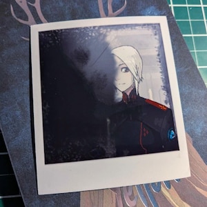 Può includere: Una stampa in stile polaroid con un personaggio anime disegnato digitalmente, con capelli bianchi e un'uniforme nera con dettagli rossi. La stampa è posta su uno sfondo blu scuro con un motivo di corna di cervo.