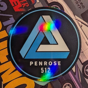 Könnte beinhalten: Ein kreisförmiger holografischer Aufkleber mit schwarzem Hintergrund. Der Aufkleber zeigt ein blau-weißes Dreieck-Design mit dem Text "PENROSE 512" in Weiß. Der Aufkleber befindet sich auf einem Magazin mit japanischem Text und Autoillustrationen.