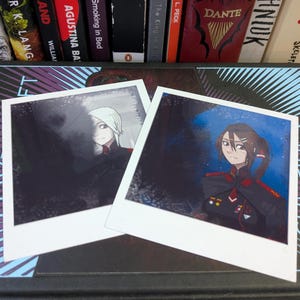 Könnte beinhalten: Zwei quadratische Fotoabzüge mit Anime-Charakterillustrationen. Der linke Abzug zeigt eine Figur mit weißem Haar in einer dunklen Uniform. Der rechte Abzug zeigt eine Figur mit braunem Haar in einer dunklen Uniform mit roten Akzenten.