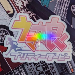 May include: A holographic sticker with Japanese text and a running figure silhouette. The sticker has a pink and purple gradient with a rainbow effect. The text includes the words "Pretty Derby".