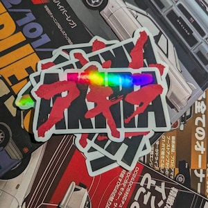 Puede incluir: Una pila de pegatinas con texto japonés rojo y una franja holográfica de arcoíris. Las pegatinas están sobre un fondo de revistas de coches con texto japonés e imágenes de coches.