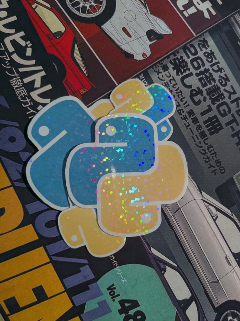 以下が含まれることがあります： 青と黄色のPythonロゴステッカーの山、ホログラフィックグリッター効果付き。ステッカーは、日本語のテキストと車のイラストが描かれた雑誌の上にあります。