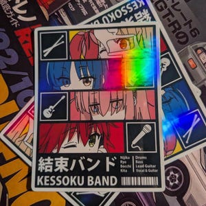 May include: A holographic sticker featuring anime-style character portraits in a grid pattern. The sticker has the text "Kessoku Band" in English and Japanese, with band member names and instruments listed below. The background includes car-related magazines.