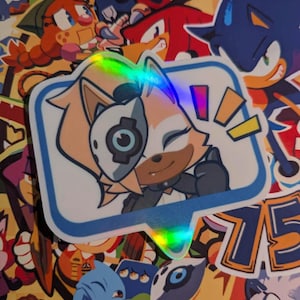May include: A sticker featuring a cartoon character with orange fur and a robotic eye, set within a blue speech bubble. The sticker has a holographic effect and is surrounded by other colorful cartoon stickers.