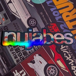 May include: A holographic sticker with the word "nubabes" in a colorful, stylized font. The sticker has a rainbow sheen and is set against a backdrop of car-themed magazine covers with Japanese text.