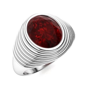 May include: A silver ring featuring a large, oval-cut, deep red gemstone. The ring's band has a layered, ridged design that surrounds the gemstone. The ring is set against a white background, highlighting its polished metal and the gemstone's brilliance.