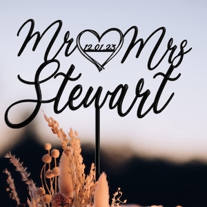 May include: A black cake topper featuring the text Mr & Mrs Stewart in a cursive script font, with a heart shape containing the date 12.01.23 positioned between the words. The topper is inserted into a white frosted cake decorated with dried flowers.