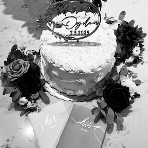 Pu&ograve; includere: Una torta nuziale bianca con glassa decorativa, sormontata da un cartello circolare con la scritta "Ogden 2.5.2025". La torta &egrave; circondata da fiori rosso scuro e bianchi. Due piccoli libri, uno con la scritta "her vows" e l'altro "his vows", sono davanti alla torta.