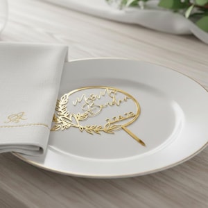 Pu&ograve; includere: Un cake topper dorato con i nomi "M&M Sandra" e la data "7.4.22" &egrave; posto su un piatto bianco. A sinistra c'&egrave; un tovagliolo bianco con iniziali ricamate in oro e un bordo dorato. Il topper &egrave; circondato da un motivo floreale.