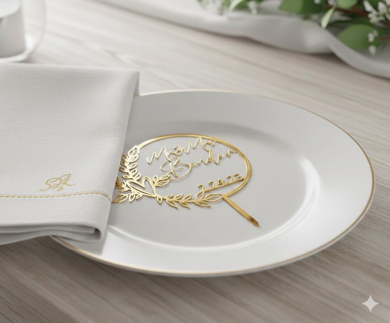 Pu&ograve; includere: Un cake topper dorato con i nomi "M&M Sandra" e la data "7.4.22" &egrave; posto su un piatto bianco. A sinistra c'&egrave; un tovagliolo bianco con iniziali ricamate in oro e un bordo dorato. Il topper &egrave; circondato da un motivo floreale.