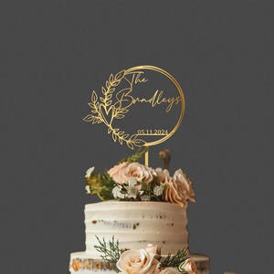 Pu&ograve; includere: Un cake topper dorato con la scritta "The Bradleys" e la data 05.11.2024, circondato da una ghirlanda floreale. Il topper &egrave; posto su una torta a pi&ugrave; strati decorata con fiori.