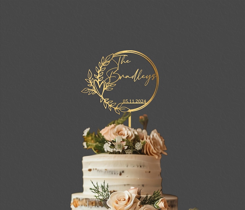 Pu&ograve; includere: Un cake topper dorato con la scritta "The Bradleys" e la data 05.11.2024, circondato da una ghirlanda floreale. Il topper &egrave; posto su una torta a pi&ugrave; strati decorata con fiori.