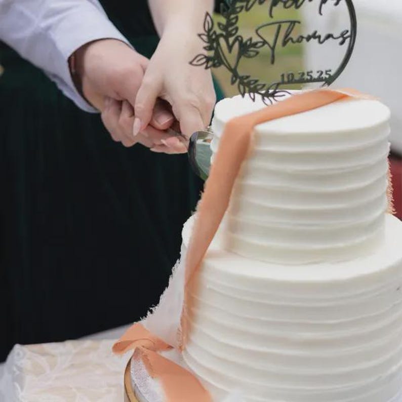 Pu&ograve; includere: Una torta nuziale bianca a due piani con glassa strutturata. Un topper decorativo con i nomi "Mr. & Mrs. Thomas" e la data "10.25.25" &egrave; in cima. Un nastro color pesca scende lungo il lato.