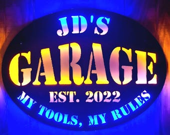 May include: A black oval metal sign with white lettering that reads "JD's Garage Est. 2022 My Tools, My Rules". The sign is illuminated with blue and yellow lights.