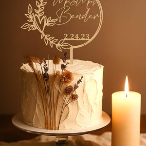 Pu&ograve; includere: Una torta nuziale glassata bianca con fiori secchi e un cake topper in legno con la scritta "Mr & Mrs Bender" e la data 2.24.23. Una candela accesa &egrave; a destra della torta.