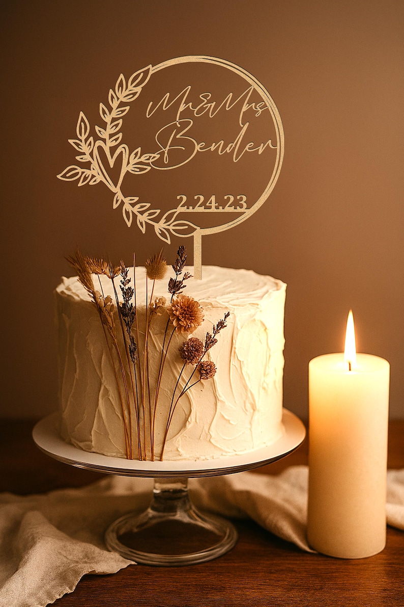 Pu&ograve; includere: Una torta nuziale glassata bianca con fiori secchi e un cake topper in legno con la scritta "Mr & Mrs Bender" e la data 2.24.23. Una candela accesa &egrave; a destra della torta.
