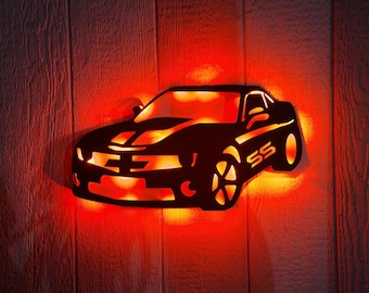 May include: A black metal silhouette of a Chevrolet Camaro SS with red LED lights. The car is mounted on a wooden wall.