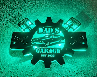 May include: A black metal sign with a gear design, two wrenches, and two pistons. The sign says "Dad's Garage Est. 2022" and features a silhouette of a classic car.