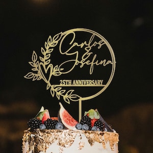 May include: A gold-colored cake topper with the names "Carlos & Josefina" and "25TH ANNIVERSARY" in script font. The topper is surrounded by a floral design and is placed on top of a cake decorated with fruit and gold leaf.