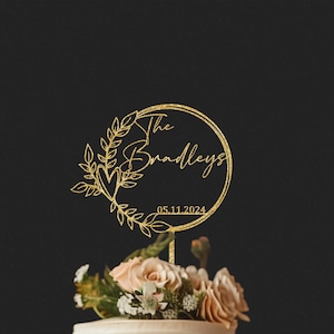 Pu&ograve; includere: Un cake topper dorato glitterato con le parole "The Bradleys" e la data 05.11.2024 all'interno di un design a ghirlanda circolare. Il topper &egrave; posto sopra una torta decorata con fiori rosa chiaro e bianchi.
