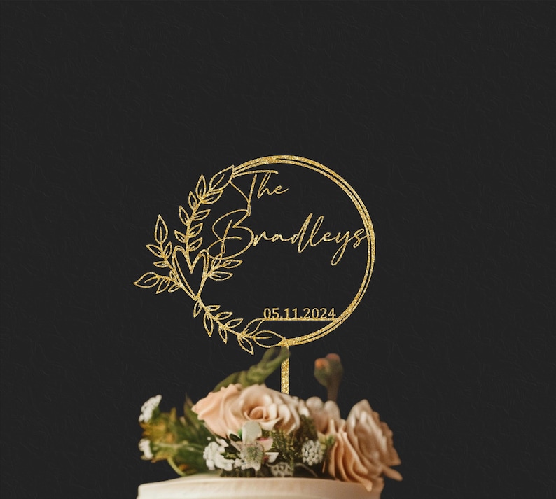 Pu&ograve; includere: Un cake topper dorato glitterato con le parole "The Bradleys" e la data 05.11.2024 all'interno di un design a ghirlanda circolare. Il topper &egrave; posto sopra una torta decorata con fiori rosa chiaro e bianchi.