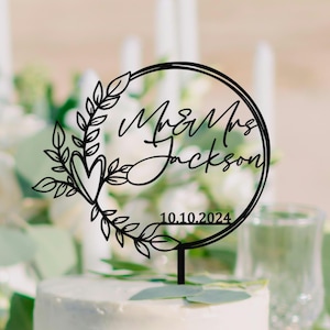 May include: Black laser-cut cake topper with the text "Mr & Mrs Jackson" and the date "10.10.2024". The topper features a heart and leaf design within a circular frame, perfect for a wedding cake.