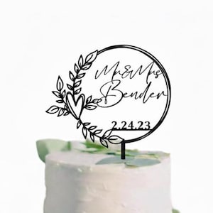 Pu&ograve; includere: Topper per torta nero tagliato al laser con la scritta "Mr & Mrs Bender" e la data "2.24.23" all'interno di una cornice circolare. La cornice &egrave; decorata con foglie e un cuore. Il topper &egrave; posto sopra una torta bianca.