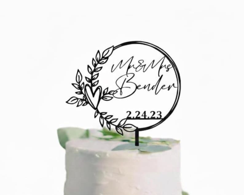 Pu&ograve; includere: Topper per torta nero tagliato al laser con la scritta "Mr & Mrs Bender" e la data "2.24.23" all'interno di una cornice circolare. La cornice &egrave; decorata con foglie e un cuore. Il topper &egrave; posto sopra una torta bianca.
