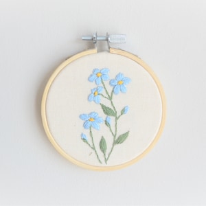 May include: A small, round embroidery hoop featuring a floral design. The artwork showcases light blue flowers with yellow centers and green leaves, stitched on a cream-colored fabric. The hoop is made of natural wood.