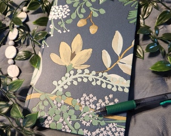 May include: A dark blue journal with a floral design featuring yellow, green, and white flowers. The journal is lying on a gray surface with green foliage.