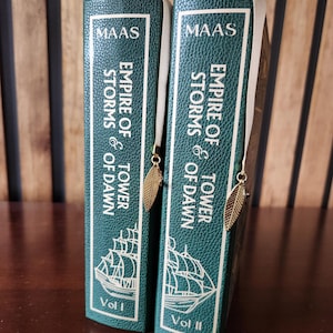 May include: Two hardcover books, "Empire of Storms & Tower of Dawn," in teal green with white lettering and a ship illustration. Each book has a cream-colored ribbon bookmark with a gold leaf charm. The author's name, MAAS, is at the top.