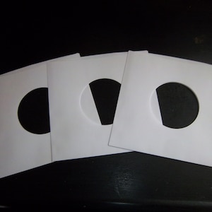 May include: Three white paper record sleeves with a round hole in the center. The sleeves are designed to protect vinyl records.