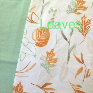 May include: A white fabric featuring a repeating pattern of orange and light green leaves, stems, and wheat stalks. The word Leaves is printed in light green text across the center of the fabric, which is positioned next to a solid light green fabric.