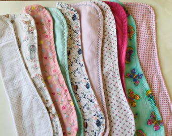 May include: A collection of eight rectangular burp cloths with rounded corners, featuring various patterns including deer, floral prints, butterflies, pink gingham, solid pink, and light blue, all finished with decorative stitched edges.