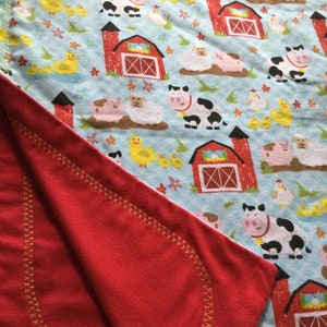May include: A red and white flannel baby blanket with a farm animal print. The blanket has a red flannel backing with a gold stitched border.