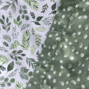 May include: A textile fabric featuring two distinct patterns: one side displays a white background with a dense print of green leaves and sprigs, while the other side is a solid sage green with white polka dots and a decorative vine-like stitch.