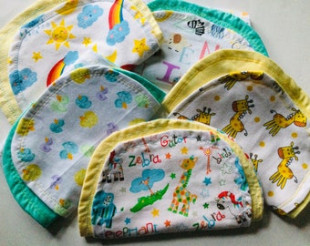 May include: A stack of six baby burp cloths with yellow trim. The cloths feature different colorful patterns including animals, rainbows, and clouds.