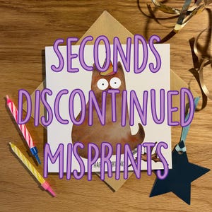 May include: A square white greeting card featuring a brown cat illustration wearing a party hat, with the text SECONDS DISCONTINUED MISPRINTS overlaid in purple. The card is accompanied by two striped birthday candles and a dark blue star ornament.