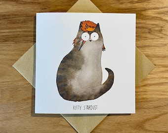 Può includere: Un biglietto d'auguri con un gatto dei cartoni animati con un ciuffo rosso e arancione, che indossa un design di trucco a forma di fulmine. Il gatto è grigio e marrone con occhi bianchi. Il testo "Kitty Stardust" è stampato sotto il gatto.