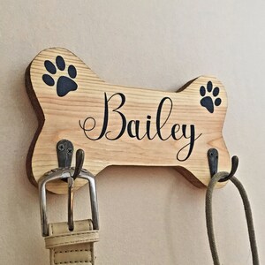 May include: Wooden dog bone-shaped leash holder with the name "Bailey" in elegant script. Features black paw prints and two metal hooks. A beige leather belt and a green rope leash hang from the hooks.