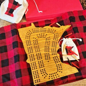 May include: A wooden game board shaped like the state of Minnesota with holes for pegs. The board is on a red and black checkered fabric. There are small bags with red and black checkered fabric and wooden pegs.