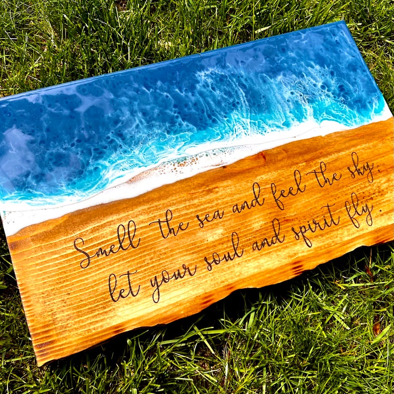 May include: A wooden sign with a blue and white epoxy resin ocean wave design. The sign has the text "Smell the sea and feel the sky, let your soul and spirit fly."